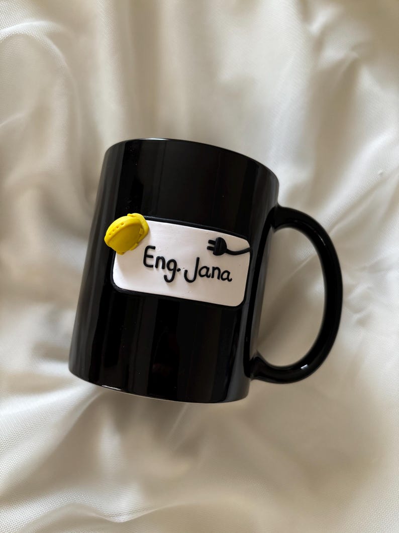 May include: A glossy black ceramic mug featuring a white rectangular plaque on the front. The plaque displays the text Eng. Jana in black script, accompanied by a small yellow hard hat icon and a black electrical plug icon attached to the text.