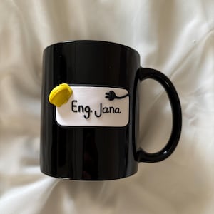 May include: A black ceramic mug featuring a white rectangular plaque with the text Eng. Jana in black script. The plaque is decorated with a small yellow hard hat and a black electrical plug icon attached to the right side of the text.