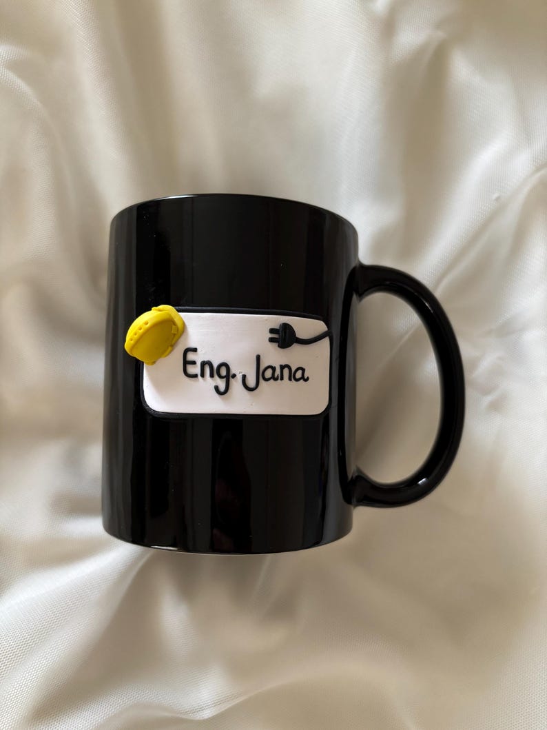 May include: A black ceramic mug featuring a white rectangular plaque with the text Eng. Jana in black script. The plaque is decorated with a small yellow hard hat and a black electrical plug icon attached to the right side of the text.