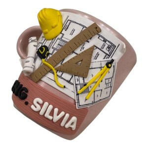 Puede incluir: Una taza de cer&aacute;mica rosa claro con un dise&ntilde;o en relieve 3D de un casco de seguridad amarillo, un plano enrollado, una regla marr&oacute;n, un tri&aacute;ngulo marr&oacute;n, una cinta m&eacute;trica amarilla y un comp&aacute;s amarillo. La taza muestra el texto ENG. SILVIA en letras blancas.