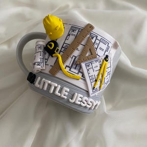 Puede incluir: Una taza de cer&aacute;mica gris decorada con elementos arquitect&oacute;nicos tridimensionales que incluyen un casco amarillo, una regla marr&oacute;n, una cinta m&eacute;trica amarilla, una escuadra y un comp&aacute;s. La taza muestra planos y el texto LITTLE JESSY.