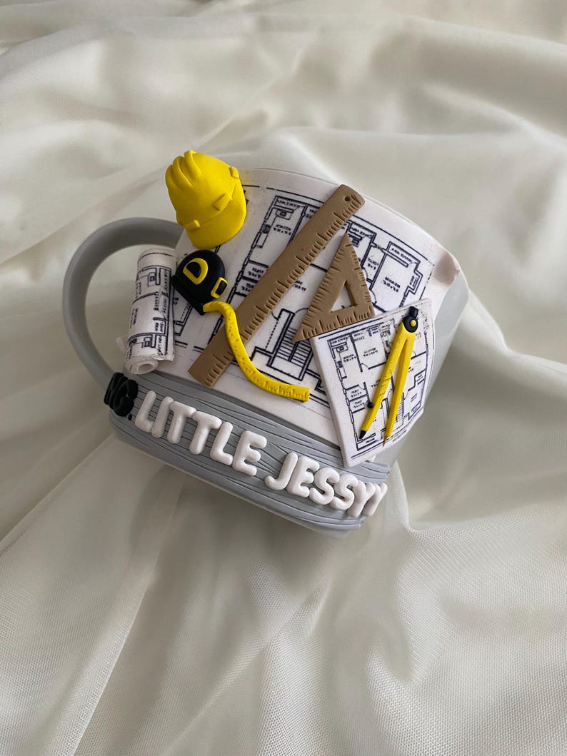 Puede incluir: Una taza de cer&aacute;mica gris decorada con elementos arquitect&oacute;nicos tridimensionales que incluyen un casco amarillo, una regla marr&oacute;n, una cinta m&eacute;trica amarilla, una escuadra y un comp&aacute;s. La taza muestra planos y el texto LITTLE JESSY.