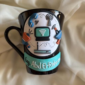 Könnte beinhalten: Schwarze Keramik-Tasse mit herzförmigem Henkel. Die Tasse hat einen weißen Kreis mit Kommunikationssymbolen, darunter ein Telefon, eine Antenne und ein Computerbildschirm. Ein türkisfarbenes Band mit dem Text "Eng. Abd El-Rahman" umschließt die Tasse.