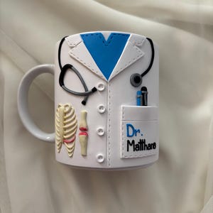 Peut inclure: Mug en céramique blanche décoré d'un design de blouse de médecin, comprenant un col bleu, un stéthoscope et une poche avec le texte "Dr. Matthane". La tasse présente également une illustration de cage thoracique et d'articulation du genou.
