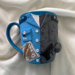 May include: A ceramic mug with a blue handle and a design split down the middle. One side is light blue with a pocket and the text "Eng. Nasr" and a blueprint. The other side is grey with a gun and camouflage pattern.