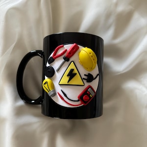 May include: A black ceramic mug featuring a circular white emblem decorated with three-dimensional miniature electrical tools including red pliers, a screwdriver, a yellow hard hat, a light bulb, a multimeter, and a yellow triangle with a black lightning bolt.