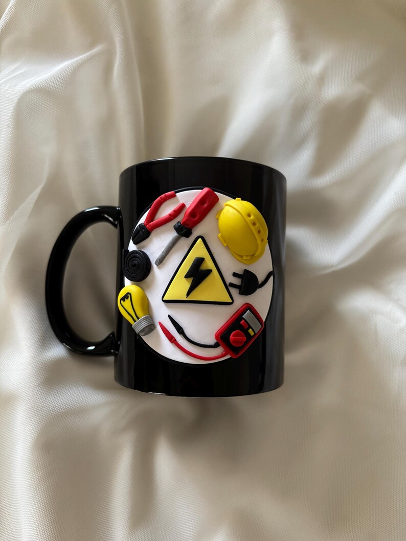 May include: A black ceramic mug featuring a circular white emblem decorated with three-dimensional miniature electrical tools including red pliers, a screwdriver, a yellow hard hat, a light bulb, a multimeter, and a yellow triangle with a black lightning bolt.