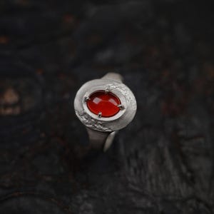 May include: A silver ring with a large, oval, red gemstone. The gemstone is set in a textured, circular bezel with small, silver prongs. The ring has a simple, smooth band.