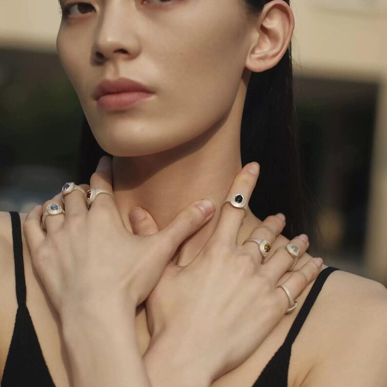 Puede incluir: Anillos de plata con piedras preciosas de varios colores. Los anillos se llevan en varios dedos. Las piedras preciosas incluyen piedras azules, rojas, amarillas y negras. La modelo lleva una camiseta sin mangas negra.