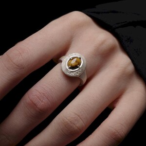 Puede incluir: Un anillo de plata con una piedra preciosa de ojo de tigre ovalada. El anillo tiene una banda blanca texturizada y un engaste decorativo alrededor de la piedra. El anillo est&aacute; en un dedo sobre un fondo oscuro.