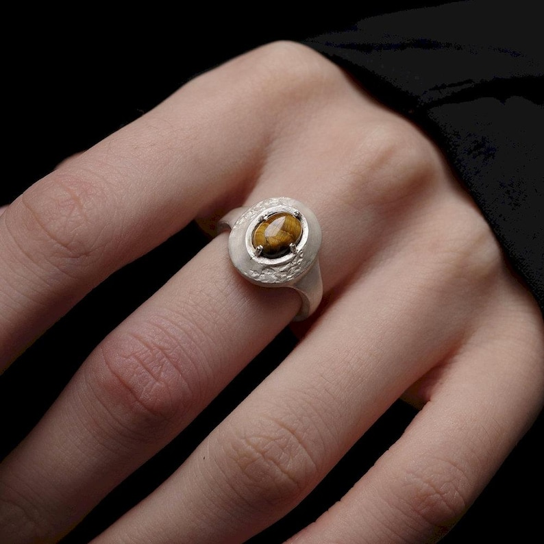 Puede incluir: Un anillo de plata con una piedra preciosa de ojo de tigre ovalada. El anillo tiene una banda blanca texturizada y un engaste decorativo alrededor de la piedra. El anillo est&aacute; en un dedo sobre un fondo oscuro.