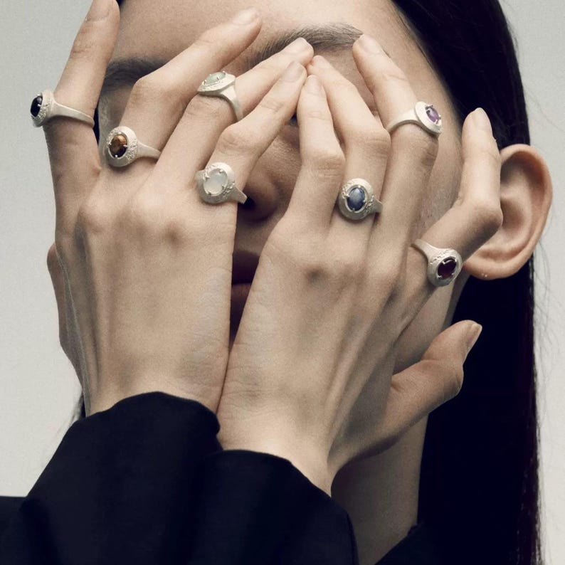 Puede incluir: Una colecci&oacute;n de anillos de plata, cada uno con una piedra preciosa diferente. Los anillos se muestran en varios dedos, mostrando varias piedras de corte ovalado y redondo en tonos negros, marrones, blancos, azules y rojos. Los anillos tienen un dise&ntilde;o simple y elegante.