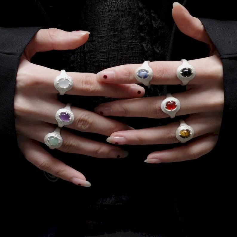 Puede incluir: Una colecci&oacute;n de anillos de plata, cada uno con una piedra preciosa de diferente color. Las piedras preciosas incluyen blanco, azul, negro, rojo, morado, verde y amarillo. Los anillos se muestran sobre un fondo oscuro.