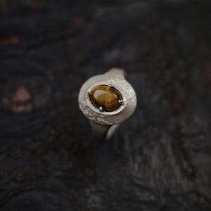 Puede incluir: Un anillo de plata con una gran piedra ojo de tigre ovalada. La piedra es de color marr&oacute;n c&aacute;lido, y el anillo tiene un dise&ntilde;o texturizado y org&aacute;nico. El anillo se muestra sobre un fondo oscuro.