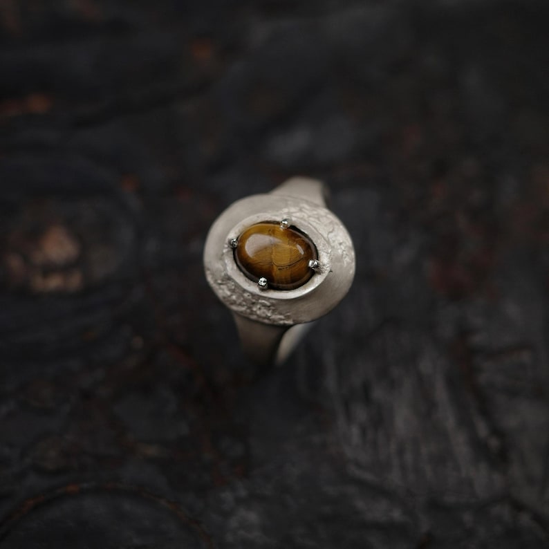 Puede incluir: Un anillo de plata con una gran piedra ojo de tigre ovalada. La piedra es de color marr&oacute;n c&aacute;lido, y el anillo tiene un dise&ntilde;o texturizado y org&aacute;nico. El anillo se muestra sobre un fondo oscuro.