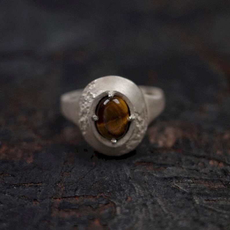 Puede incluir: Un anillo de plata con una gran piedra ojo de tigre ovalada. La piedra es de color marr&oacute;n c&aacute;lido con vetas doradas. El anillo tiene un aspecto texturizado, casi martillado, alrededor del engaste de la piedra.