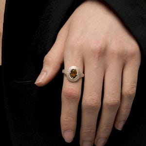 Puede incluir: Un anillo de plata con una piedra preciosa ovalada de color &aacute;mbar. El anillo tiene una banda texturizada y un engaste decorativo alrededor de la piedra. El anillo se muestra en una mano sobre un fondo negro.