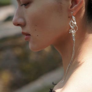 May include: A single silver earring with an abstract design. The earring has an open, web-like structure at the top, leading to a long, flowing chain. The design is intricate and modern, with organic shapes and textures. The earring is worn on the ear.