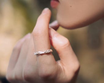 May include: A silver ring with a textured, woven design. The ring is worn on a finger, showcasing its intricate details and craftsmanship. The ring's surface reflects light, highlighting its metallic sheen. The ring is a piece of jewelry.