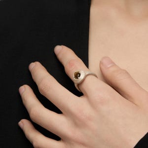 Puede incluir: Un anillo plateado con una banda texturizada y una piedra central de color &aacute;mbar en forma ovalada. El anillo se lleva en un dedo. La imagen muestra una mano y parte de una prenda negra.