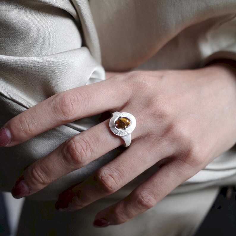 Puede incluir: Un anillo de plata con una gema redonda de color &aacute;mbar engastada en un dise&ntilde;o calado texturizado. El anillo se muestra en una mano, mostrando los detalles de la joya. El dise&ntilde;o del anillo es simple pero elegante.
