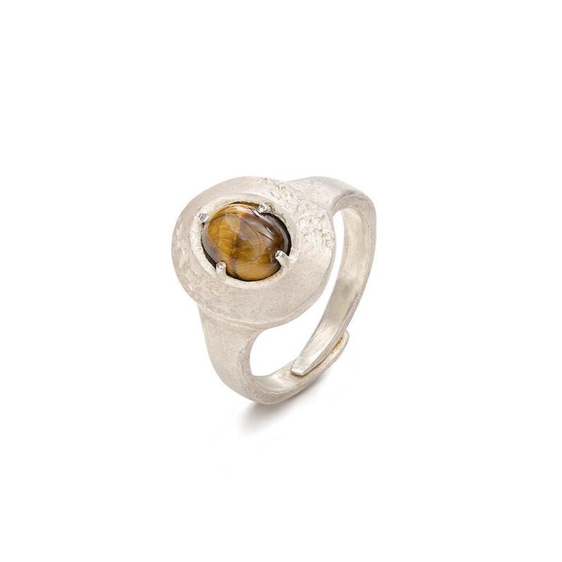 Puede incluir: Anillo de plata con un engaste redondo y texturizado. El anillo presenta una piedra ojo de tigre ovalada pulida. La banda es ajustable. El anillo est&aacute; sobre un fondo blanco.