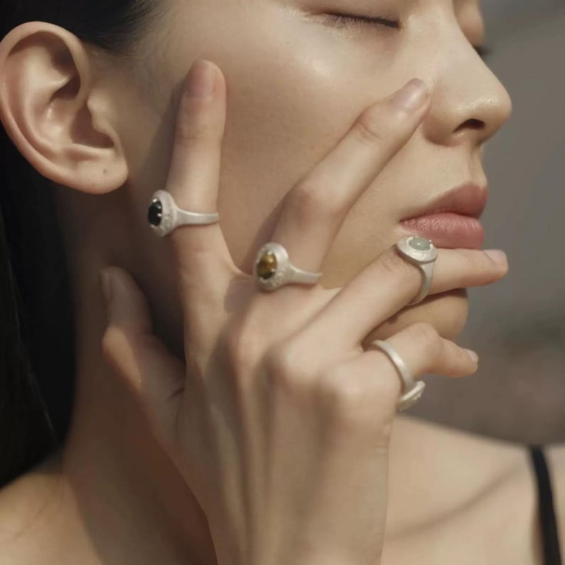 Puede incluir: Una colecci&oacute;n de anillos de plata con diferentes engastes de piedras preciosas. Los anillos presentan una gama de colores de piedras, incluyendo negro, amarillo y verde p&aacute;lido. Los anillos se muestran en los dedos, mostrando su dise&ntilde;o y detalle.