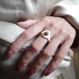 Puede incluir: Un anillo de plata con una piedra preciosa redonda de color &aacute;mbar engastada en un bisel texturizado. El anillo est&aacute; en una mano con u&ntilde;as pintadas, sobre un fondo suave y neutro.