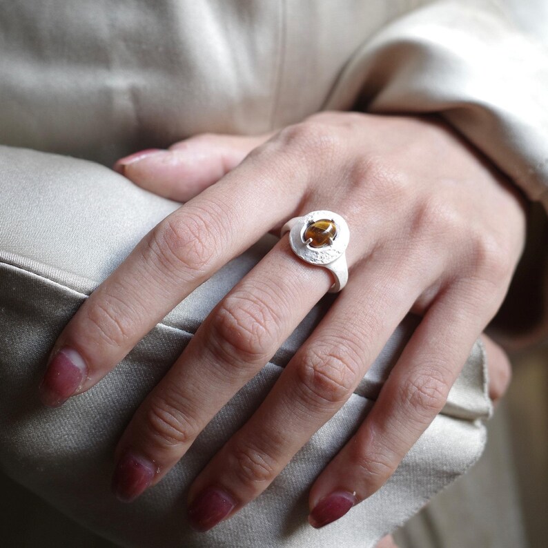 Puede incluir: Un anillo de plata con una piedra preciosa redonda de color &aacute;mbar engastada en un bisel texturizado. El anillo est&aacute; en una mano con u&ntilde;as pintadas, sobre un fondo suave y neutro.