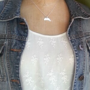May include: A silver necklace with a pendant shaped like the state of Rhode Island. The necklace is worn by a person wearing a white shirt with a floral pattern and a denim jacket.