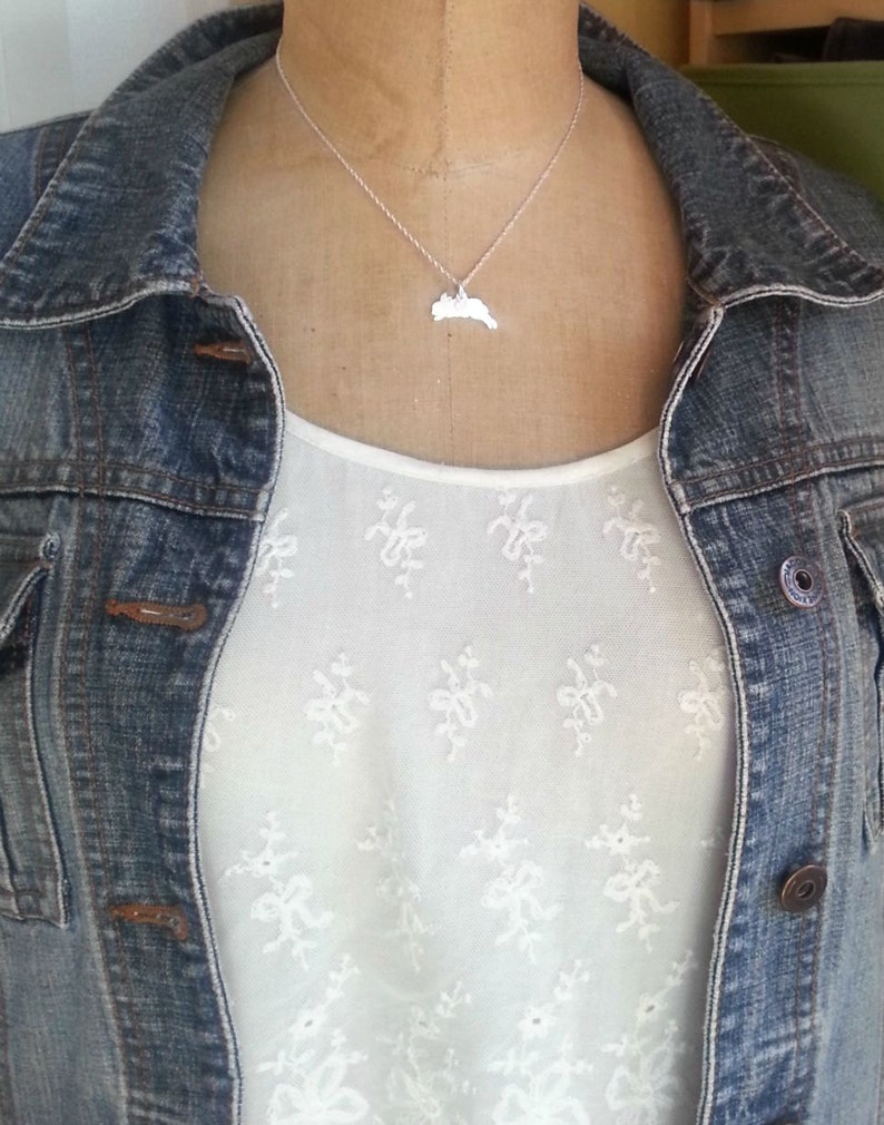 May include: A silver necklace with a pendant shaped like the state of Rhode Island. The necklace is worn by a person wearing a white shirt with a floral pattern and a denim jacket.