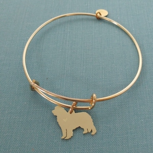 May include: Gold-colored expandable bracelet with a charm featuring a dog silhouette. The charm is a detailed outline of a dog. The bracelet is made of a thin, shiny metal and has a small, round tag attached.