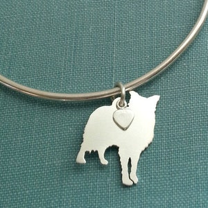 May include: A silver-colored bangle bracelet with a charm. The charm is a silhouette of a dog with a small heart charm attached. The dog charm is a light silver color and is hanging from the bracelet.