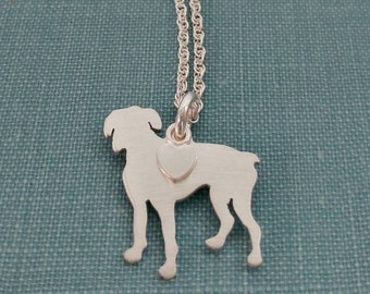 Puede incluir: Collar con colgante de perro plateado. El colgante es una silueta de un perro con un detalle de corazón, suspendido de una fina cadena. El collar se presenta sobre un fondo verde azulado. Diseño sencillo y elegante.