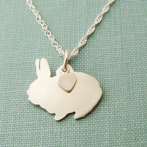 Può includere: Una collana d'argento con un ciondolo a forma di silhouette di coniglio e un piccolo ciondolo a forma di cuore. La collana è su uno sfondo blu testurizzato.