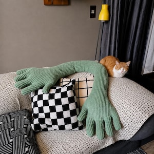 Pode incluir: Uma almofada verde em forma de braço de malha canelada com uma mão na extremidade, repousando num sofá com almofadas estampadas. Um gato ruivo está enrolado no sofá. As almofadas têm padrões a preto e branco.