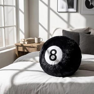 May include: A black, round cushion designed to look like an eight ball, featuring a white circle with the number 8 in the centre. The cushion is placed on a white bed. The room includes a window, a wooden bedside table, and framed art.
