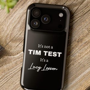 May include: Black phone case with the text "It's not a TIM TEST It's a Lucy Lesson" in white. The case has a glossy finish and a camera cutout. The phone case is on a wooden surface.