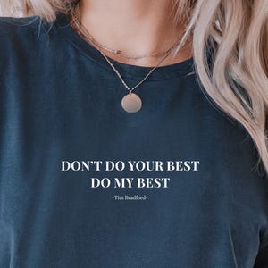 May include: Navy blue t-shirt with the text "DON'T DO YOUR BEST DO MY BEST -Tim Bradford-" in white. The shirt has short sleeves and a relaxed fit. The model is wearing a gold necklace.