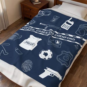 May include: A navy blue fleece blanket featuring a white line-art pattern of police-themed icons including a badge, handcuffs, a donut, a fingerprint, a handgun, and a vest. A central banner displays the text COFFEE, CASES AND CHENFORD.