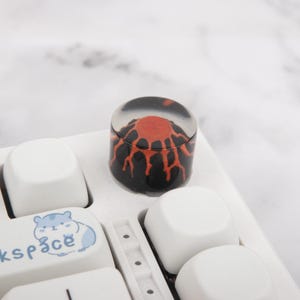 Peut inclure: Un keycap personnalisé avec un motif de volcan. Le keycap a un dessus transparent et arrondi avec un graphique de volcan rouge et noir. Il est posé sur un clavier blanc avec d'autres keycaps blancs et un keycap avec une illustration de hamster bleu.