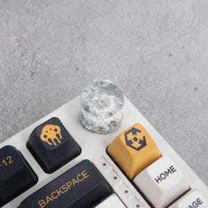 May include: A white mechanical keyboard with custom keycaps. A clear, marble-patterned keycap sits above the keyboard. Other keycaps are black and yellow with unique symbols and the word "BACKSPACE".