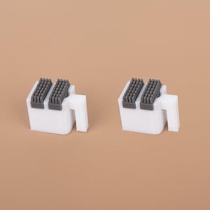 May include: Two white plastic clips, each featuring a rectangular base with a protruding side tab and two parallel dark gray rubberized grip strips on the top surface, designed for securing items with a firm hold and a clean, functional appearance.
