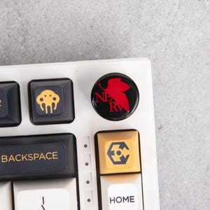 Op de afbeelding: Een close-up van een wit toetsenbord met aangepaste keycaps. Eén keycap is zwart met een rood ontwerp en de letters "NE RV". Andere keycaps zijn zwart en geel met verschillende symbolen. Het toetsenbord ligt op een grijze ondergrond.