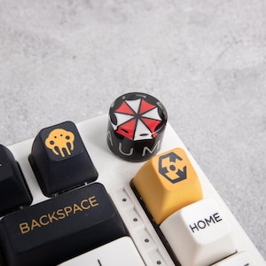 May include: A close-up of a white keyboard with custom keycaps. One keycap features a red and white umbrella logo on a black background. Other keycaps are black and yellow with various symbols and text.