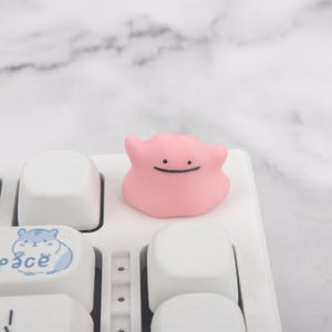 May include: A small, light pink, blob-shaped character figurine with two small protrusions on top, featuring two black dots for eyes and a simple black horizontal line for a mouth, resting on a white computer keyboard keycap.