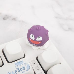 May include: A purple cartoon character keycap with a smiling face and white eyes, placed on a white keyboard. The keycap has a round body with small holes and a white, cloud-like base. The keyboard keys are white.