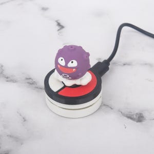 May include: A purple spherical character with a wide grin and white eyes sits atop a circular charging pad designed like a red, white, and black Poké Ball. A black charging cable extends from the side of the device, which features a textured surface.