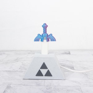 Puede incluir: Una lámpara de escritorio con forma de espada incrustada en una base de piedra. El mango de la espada tiene un acabado degradado en azul y morado, mientras que la base es gris claro con textura moteada y un símbolo triforce negro al frente.