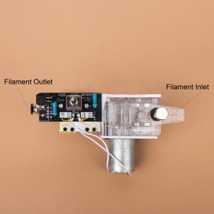 May include: A 3D printer filament extruder mechanism featuring a black circuit board with a toggle switch labelled FEED, STOP, and RETRACT. It includes a clear plastic housing, a silver cylindrical motor, and two blue resistors, with text labels Filament Outlet and Filament Inlet.